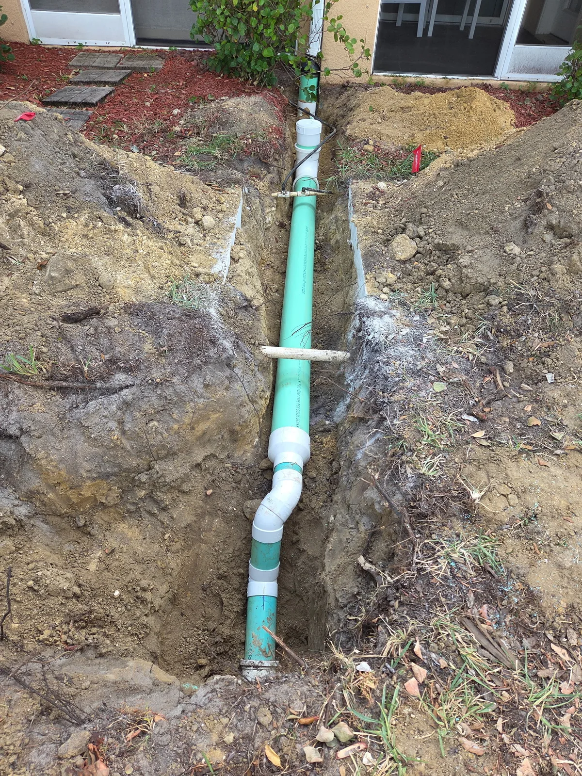 Green PVC sewer line laid in a gravel trench along the side of a house